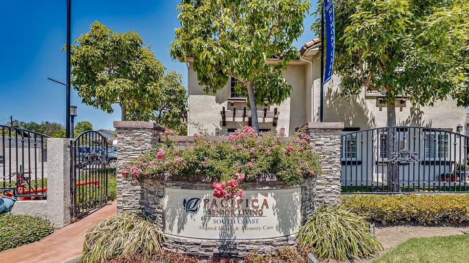 Entrance sign for Pacifica Senior Living South Coast with stone pillars, a black metal gate, flowering bushes, and trees in front of a beige building under a clear blue sky.