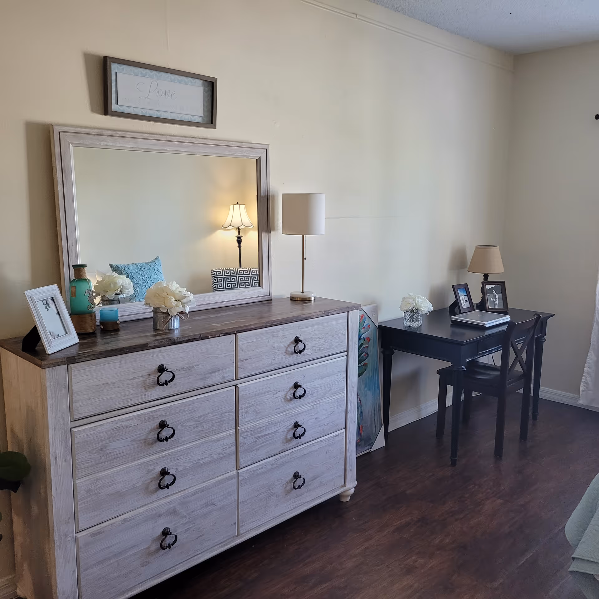 A bedroom corner with a whitewashed dresser topped by a large mirror and decorative items, and a small black desk and chair against a pale wall.
