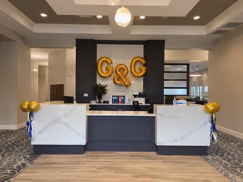 Reception desk area with a modern design featuring a marble-like countertop and dark cabinetry. Behind the desk, gold balloon letters spell out 'G&G'. The floor has a combination of wood and patterned carpet, and the space is well-lit with ceiling lights and a hanging pendant light.