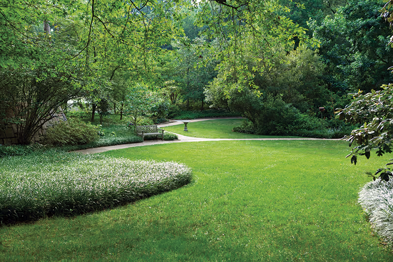 A lush green garden area with well-maintained grass, various shrubs, and trees. There is a paved pathway winding through the garden and a wooden bench placed near the path, surrounded by dense greenery.