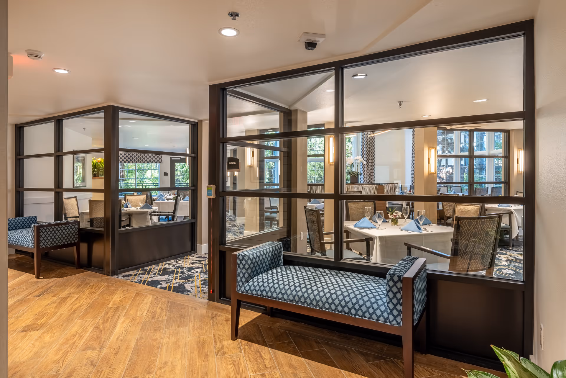 Interior view of a senior living facility showing a hallway with wooden flooring and a blue patterned bench. Through large glass windows, a dining room is visible with tables set with white tablecloths, blue napkins, glassware, and chairs. The dining room has large windows letting in natural light and decorative wall sconces.
