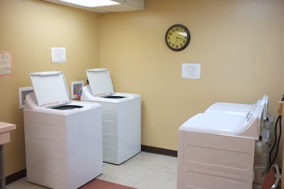 A laundry room with four white top-loading washing machines, two on the left with lids open and two on the right with lids closed. The walls are beige, and there is a round clock showing the time as 4:45. There are two printed notices taped to the wall above the machines.