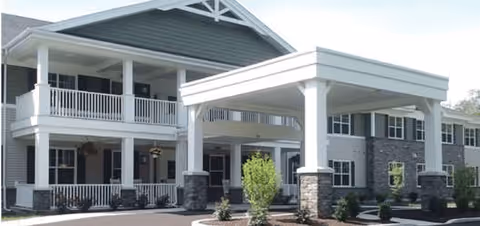 Exterior view of a two-story senior living facility building with a covered entrance supported by white columns and stone bases. The building has a gray roof, white railings on the upper balcony, and several windows. There are small shrubs and landscaping around the entrance area.