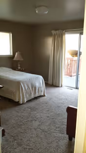 A bedroom with a single bed covered in a white bedspread, a bedside table with a lamp, a window with blinds, and a sliding glass door with curtains leading to an outdoor balcony or patio. The room has beige walls and carpeted flooring.