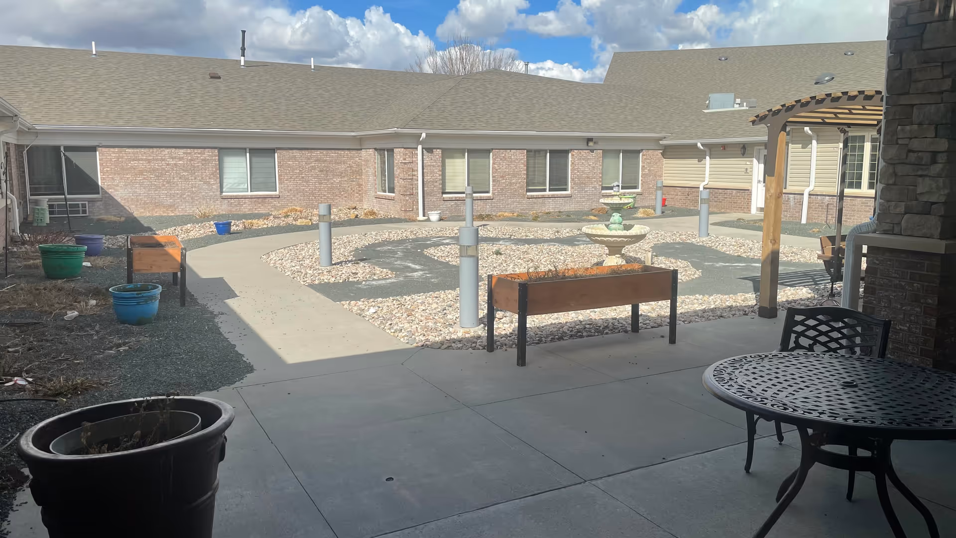 Outdoor courtyard area with a concrete pathway, decorative rocks, planters, a water fountain, and patio furniture including a round metal table and chairs. The courtyard is surrounded by a single-story brick building under a partly cloudy sky.