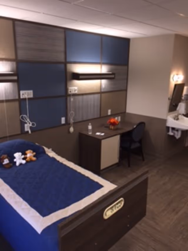 A neatly arranged bedroom in a senior living facility with a bed covered in a blue quilt and three small stuffed animals on it. The room has a modern design with a multi-colored panel wall, a desk with a chair, a small vase with flowers, and a sink with a mirror on the right side.