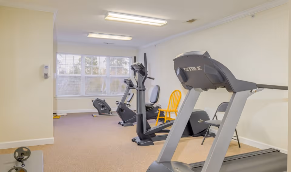 A small fitness room with exercise equipment including a treadmill, stationary bike, and elliptical machine. The room has beige walls, carpeted floor, and a large window letting in natural light. There are two chairs, one yellow wooden and one gray folding, placed near the equipment.