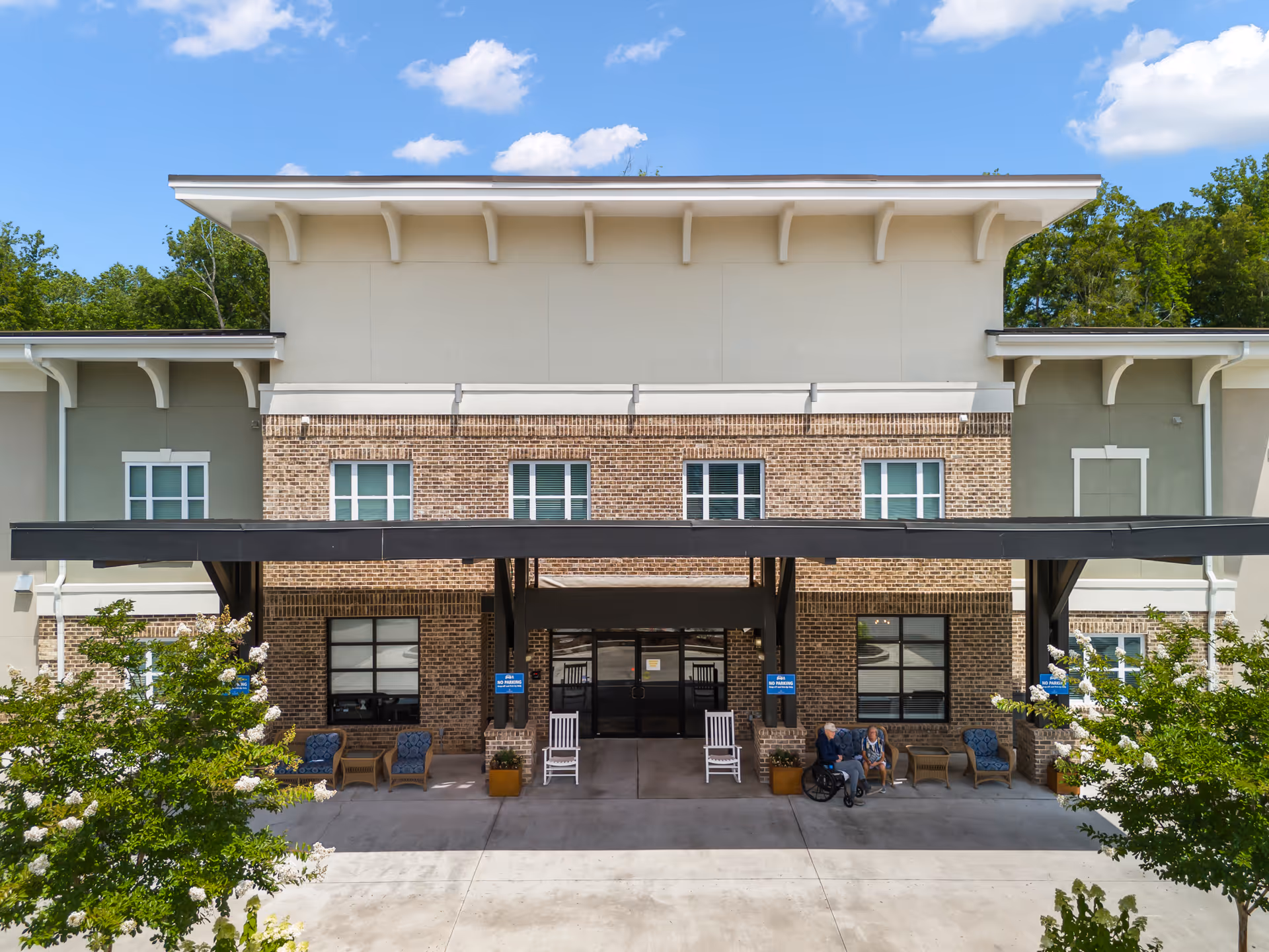 Front entrance of The Addison of Sandy Springs with a covered portico, brick facade, outdoor seating, and two people near the doorway.