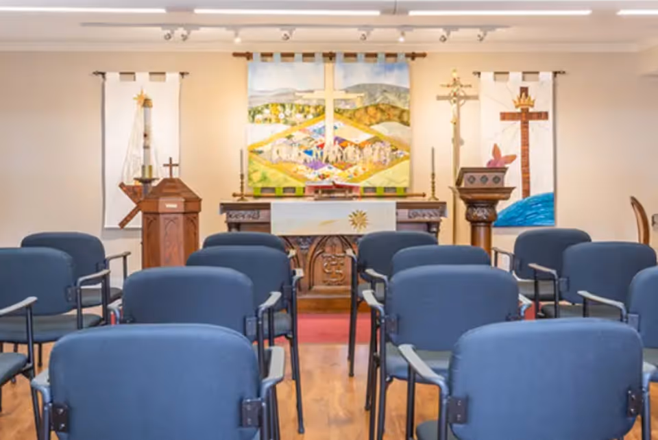 Interior view of a small chapel or worship room with rows of blue chairs facing an altar adorned with religious banners and crosses.