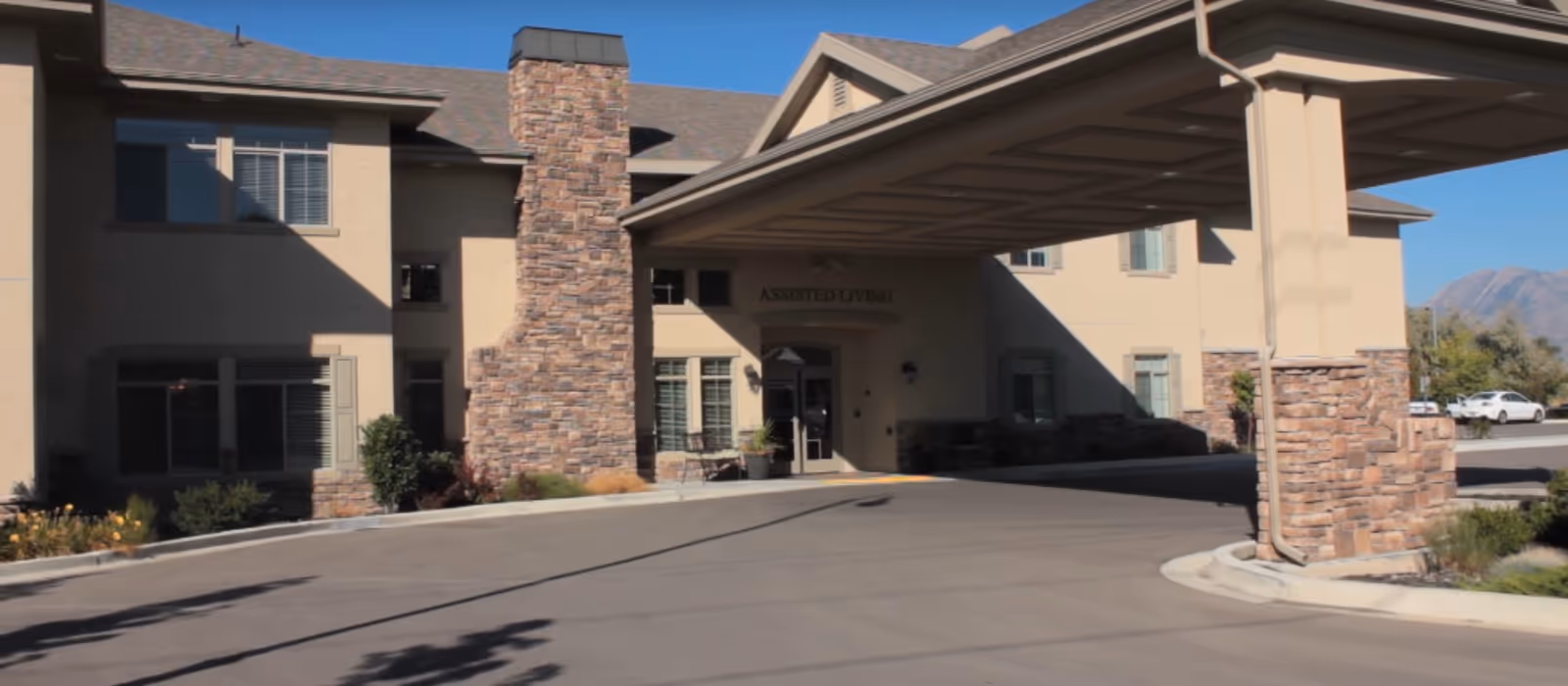 Front exterior view of Carrington Court Assisted Living and Memory Care building with a covered entrance, stone pillars, and a driveway under a clear blue sky.