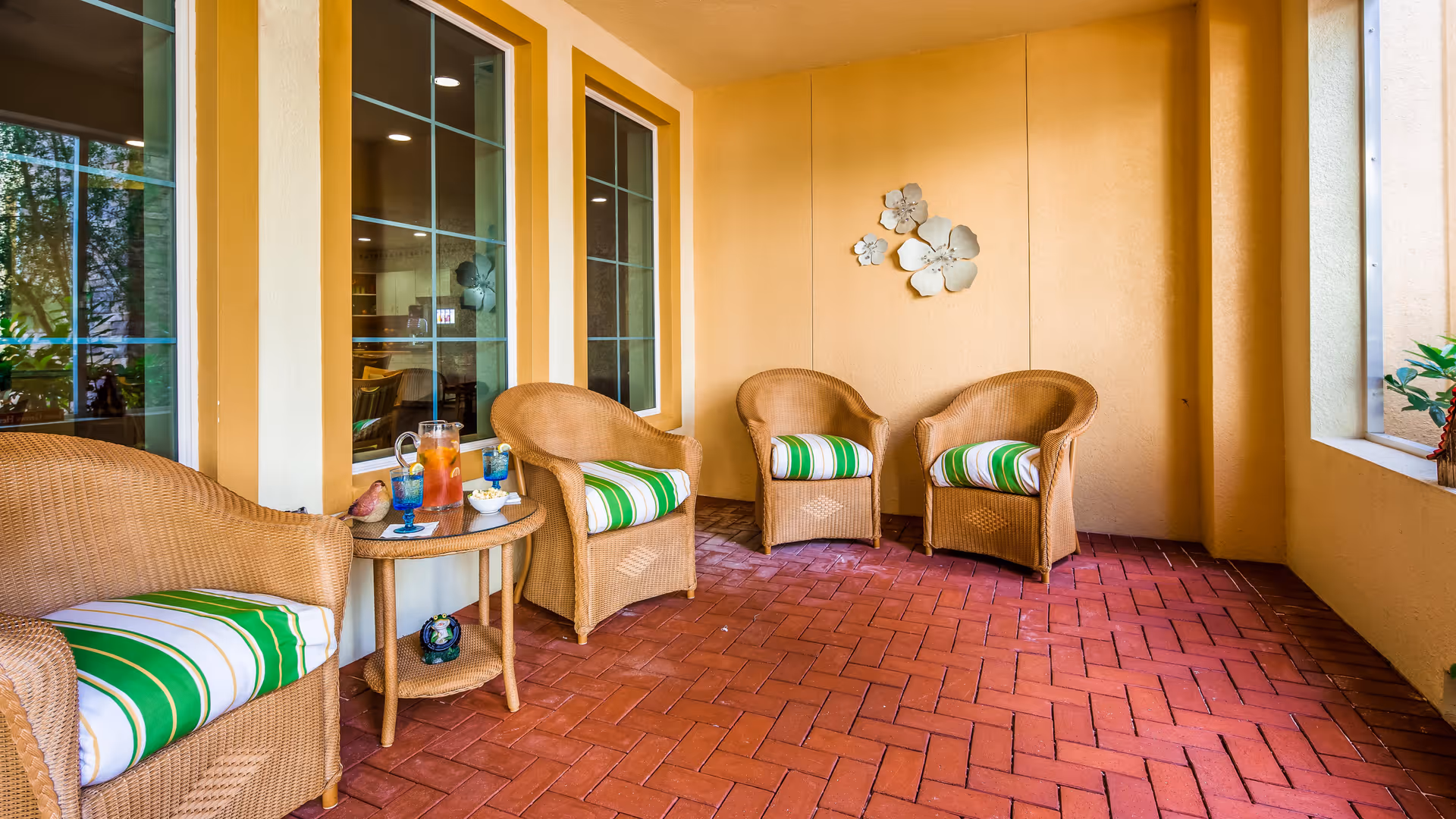 A cozy indoor seating area with four wicker chairs featuring green and white striped cushions arranged around a small wicker table. The table holds a pitcher of iced tea, two blue glasses, and a small decorative item. The walls are painted a warm beige color with three decorative flower wall hangings. Large windows look into another room, and the floor is covered with reddish-brown brick tiles.