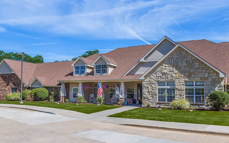 Exterior view of a single-story senior living facility building with a stone and siding facade, a brown shingled roof, and a well-maintained lawn. The entrance has a covered porch with hanging flower baskets, American flags, and outdoor seating. The sky is clear and blue.