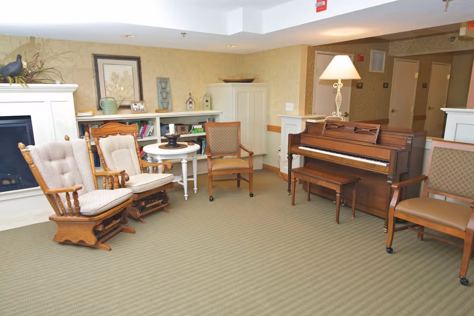 A cozy senior living common area with two cushioned wooden rocking chairs, a small round white table with a candle holder, a wooden upright piano with a bench, a floor lamp, and two cushioned armchairs. The room has beige patterned wallpaper, a carpeted floor, and a white fireplace with decorative items on the mantel and shelves.