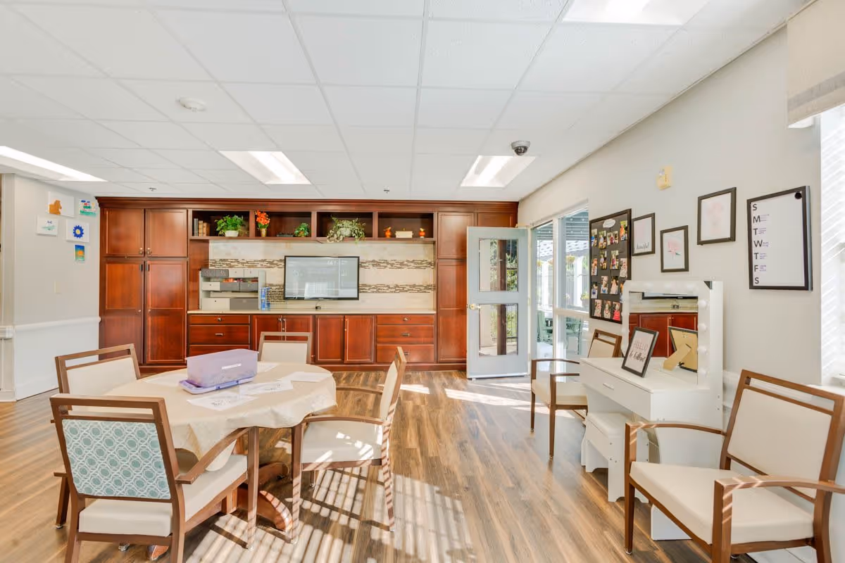 A bright and spacious room with wooden flooring featuring a round table covered with a beige tablecloth surrounded by four chairs. The back wall has built-in wooden cabinets with shelves decorated with plants and books, and a flat-screen TV mounted in the center. To the right, there is a white vanity table with a mirror and two chairs. The room has large windows allowing natural light to enter, and a door leading outside.