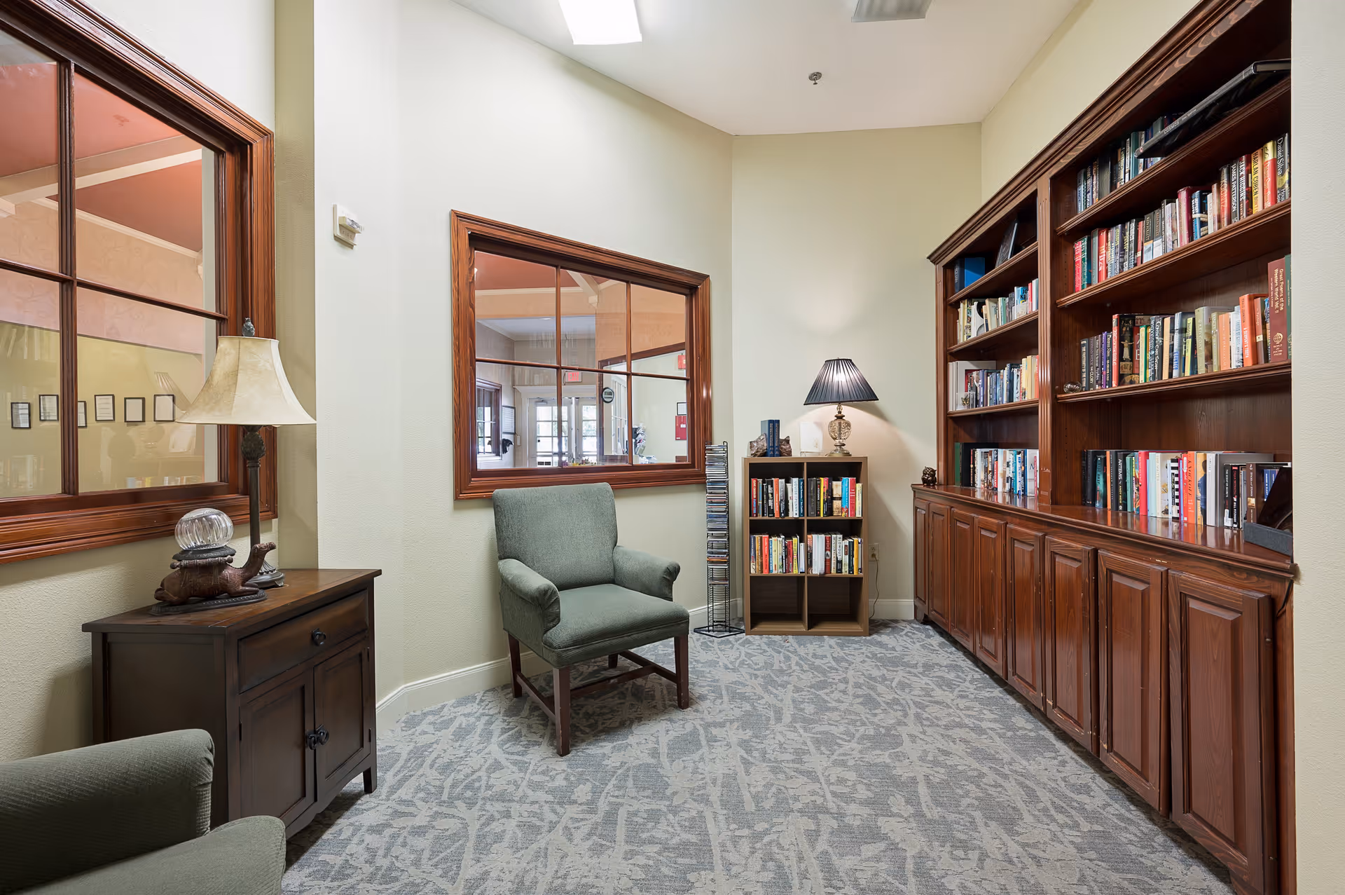 A cozy reading nook in a senior living facility featuring a green upholstered armchair, a wooden side table with a lamp and decorative items, a tall wooden bookshelf filled with books, and a smaller bookshelf with additional books and a lamp on top. The walls are light-colored and there are two large windows with wooden frames looking into another room.