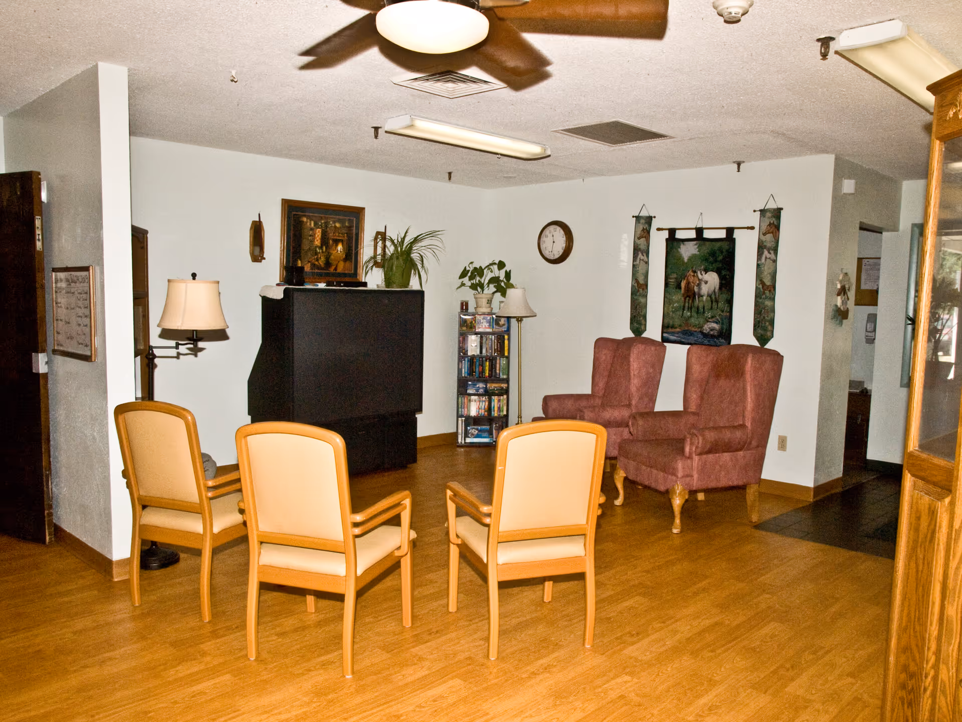 Communal sitting area with four beige chairs and two upholstered armchairs facing a large TV and shelving in a nursing home lounge.