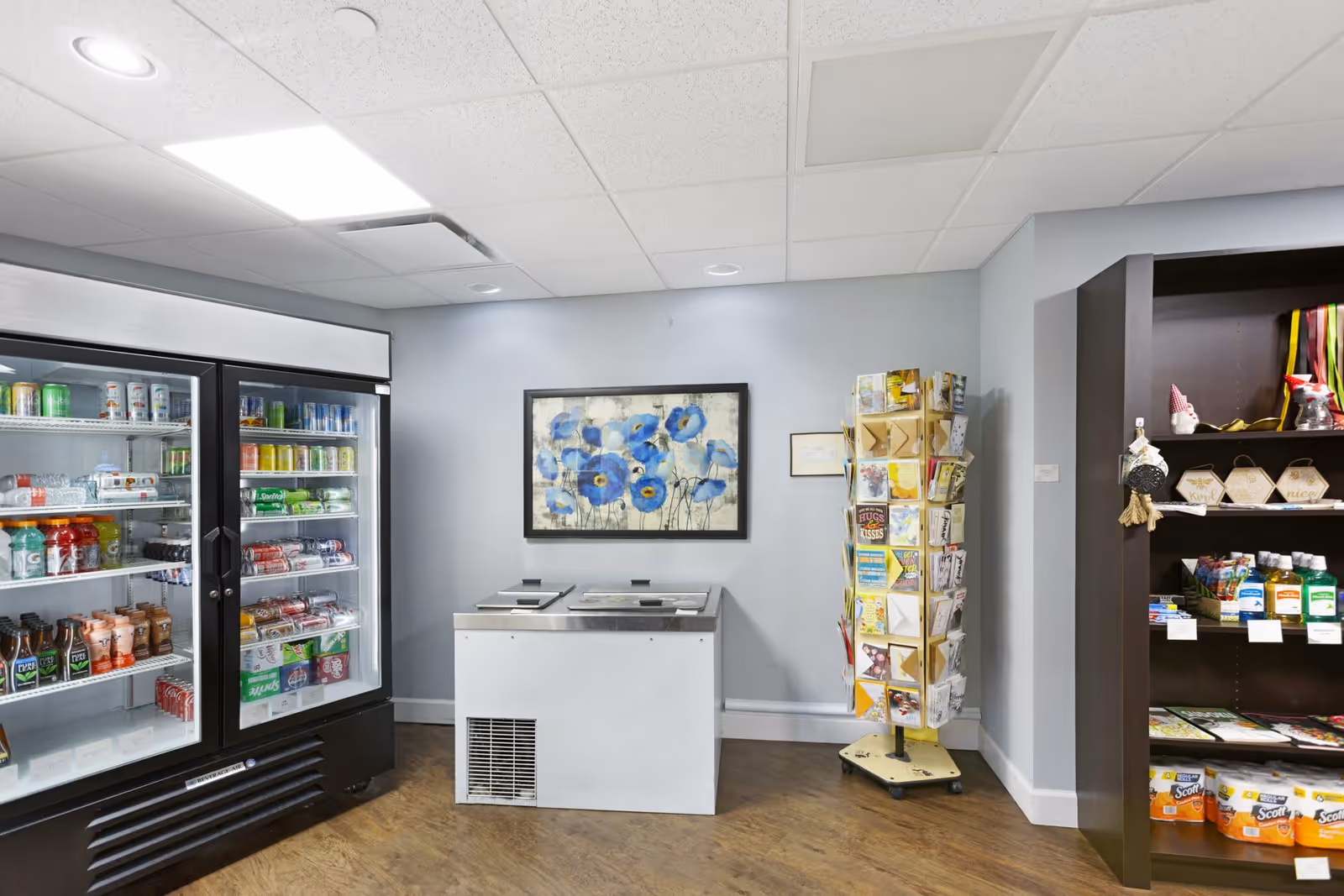 Interior view of a small convenience area in an assisted living facility featuring a glass-door refrigerator stocked with various beverages, a white freezer chest, a rotating rack filled with greeting cards, and a dark wooden shelf stocked with snacks and household items. A framed painting of blue flowers hangs on the light gray wall above the freezer.