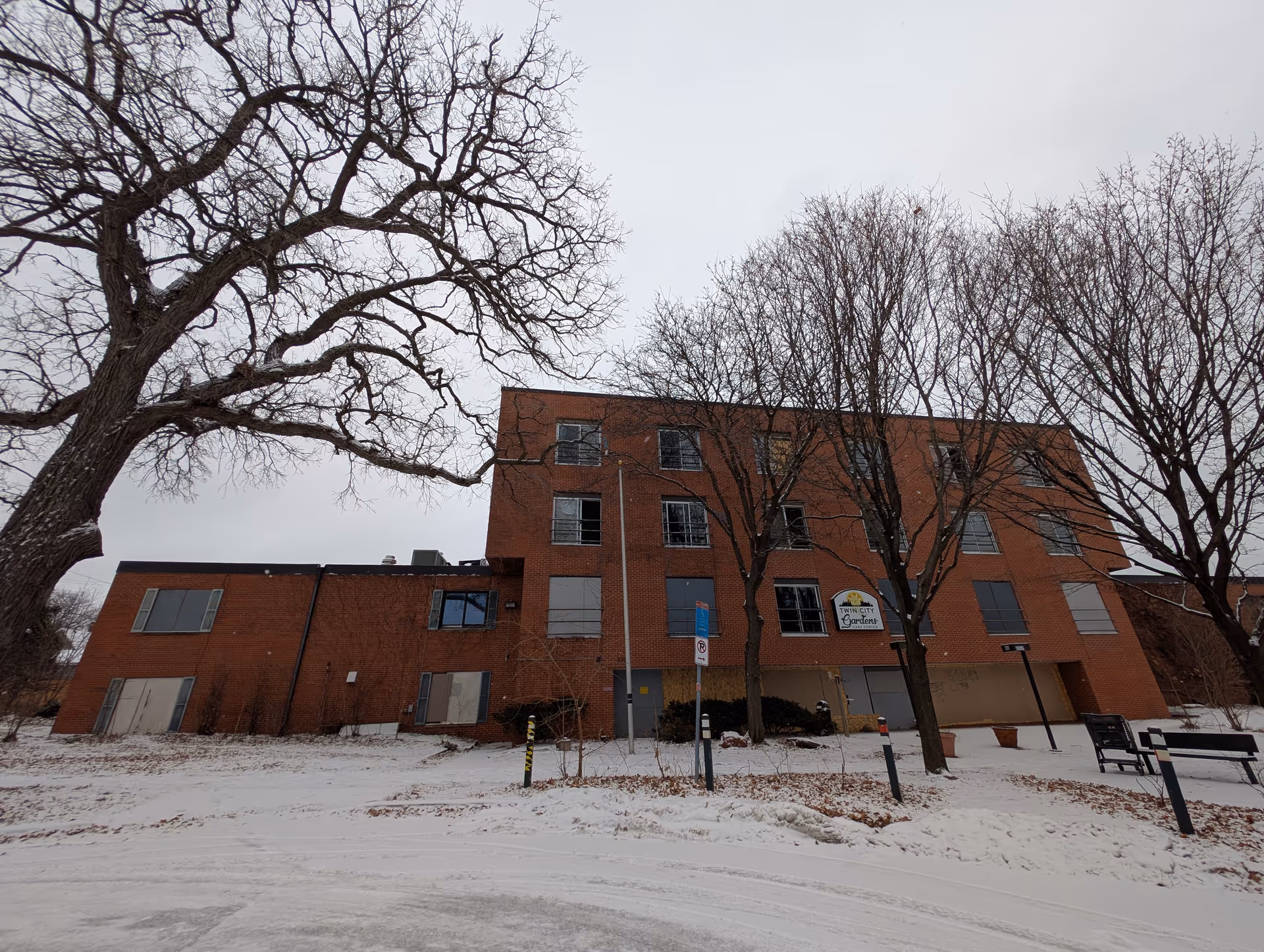 Exterior view of a three-story brick building in winter with leafless trees and snow on the ground. The building has boarded-up windows and a sign that reads 'Twin City Gardens Care Center'.