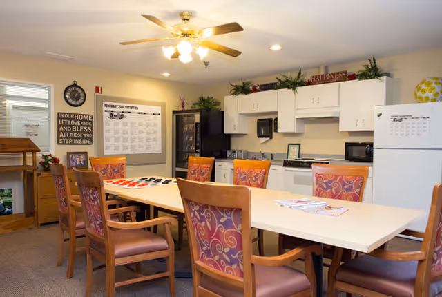 A well-lit common room with a long table surrounded by wooden chairs with patterned cushions. The room features a ceiling fan with lights, a kitchenette area with white cabinets, a refrigerator, microwave, and a stove. On the wall, there is a large calendar and a decorative sign with an inspirational quote. The room has a cozy and welcoming atmosphere.