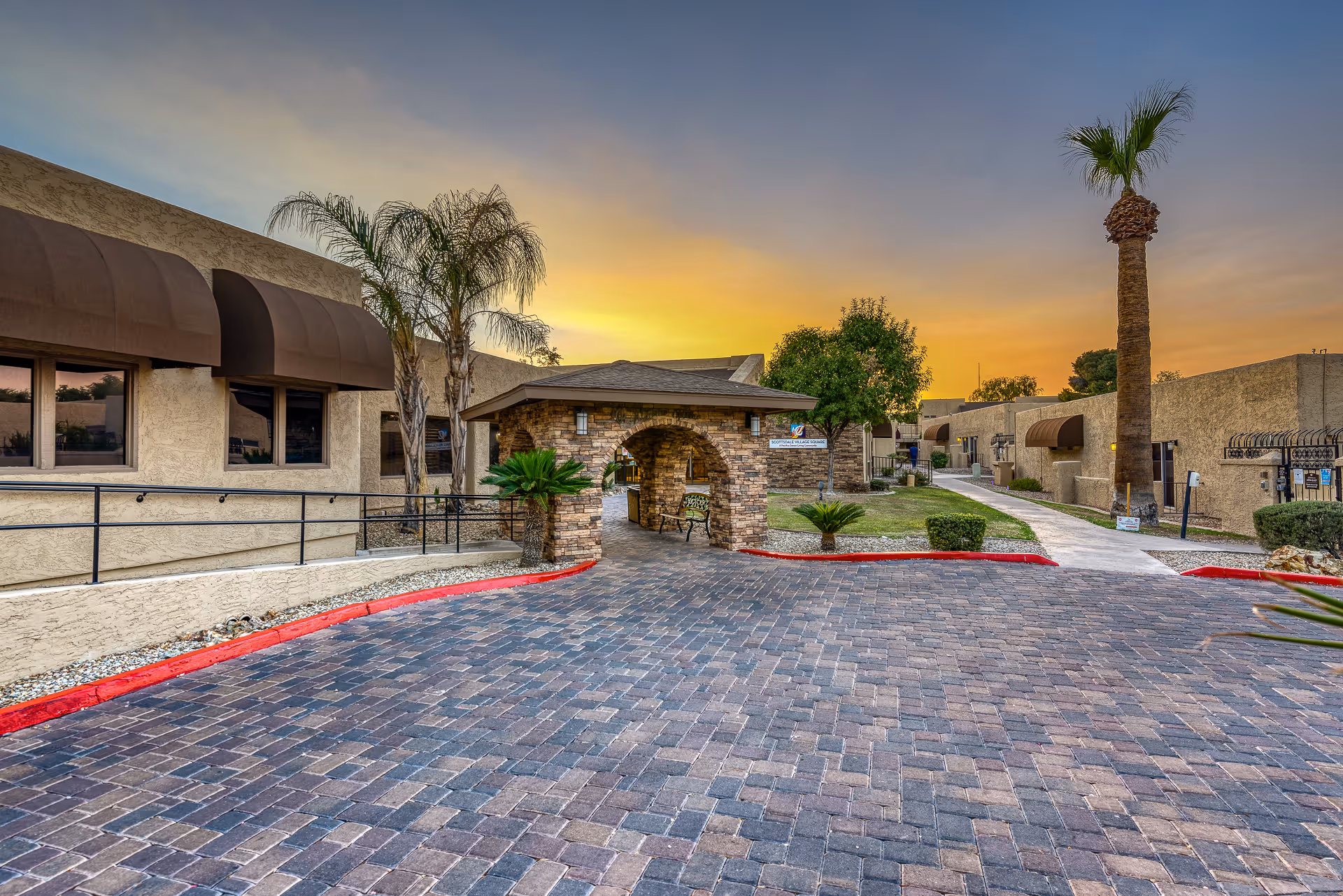 Exterior view of Scottsdale Village Square senior living facility at sunset, showing a paved driveway, a small stone archway with a bench underneath, palm trees, and beige buildings with brown awnings.