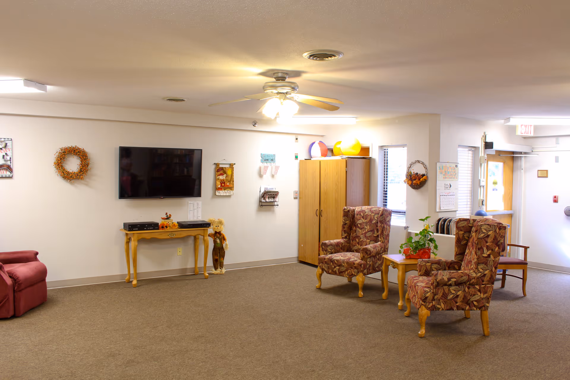 A cozy living room area in a senior living facility with two patterned armchairs and a small wooden table with a plant on it. There is a flat-screen TV mounted on the wall above a small wooden table with decorative items and a teddy bear standing beside it. The room has a ceiling fan with lights, a cabinet, and windows letting in natural light. The walls are decorated with wreaths and small wall hangings.