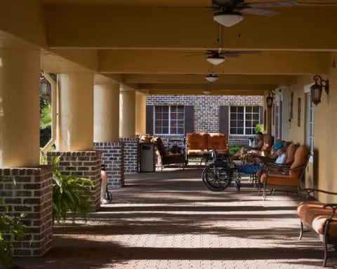 Covered outdoor veranda with columns and brick half-walls, cushioned seating, a wheelchair, ceiling fans, and potted plants.