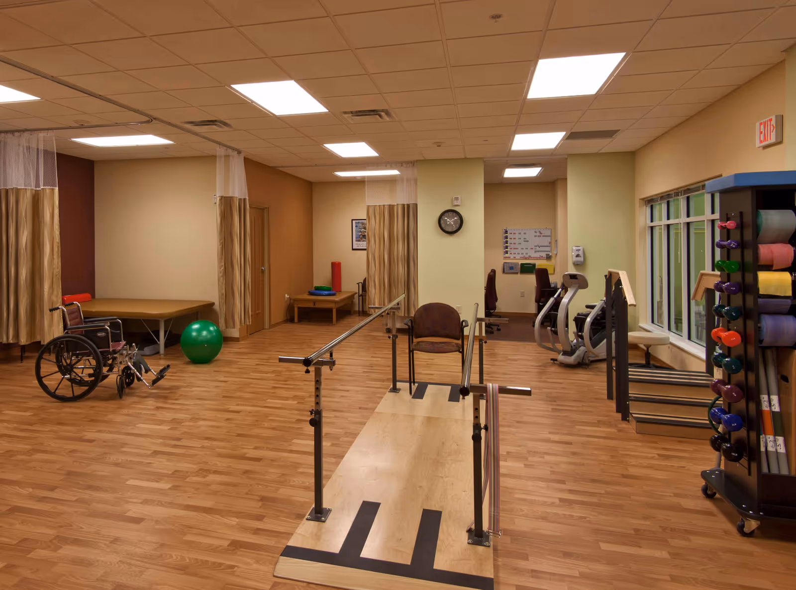 A physical therapy room with wooden flooring, parallel bars for walking exercises, a wheelchair, exercise balls, therapy tables with privacy curtains, a rack with colorful dumbbells and exercise mats, and exercise machines near a window.