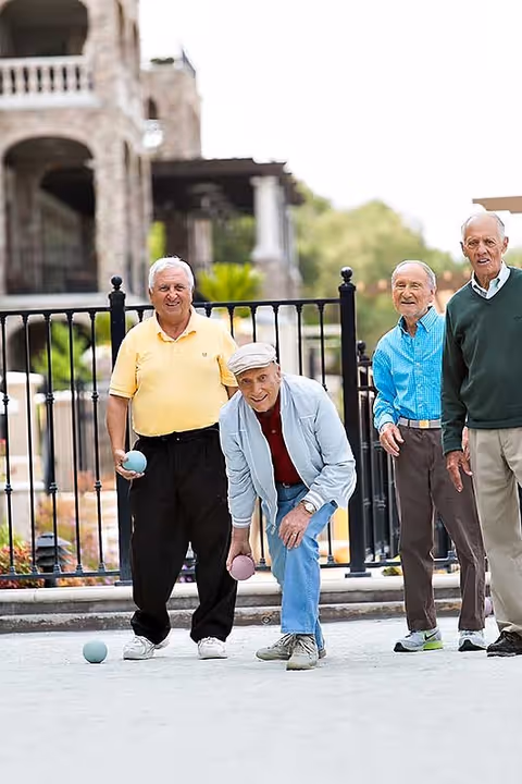 Four elderly men playing bocce ball outdoors on a paved court with a stone building and black metal fence in the background.