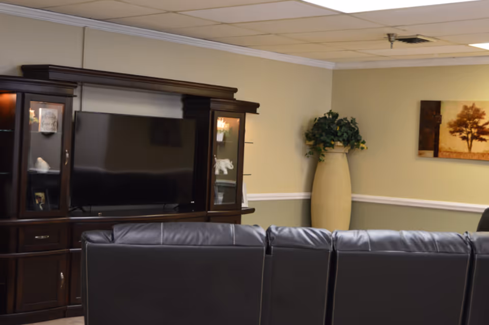 A cozy living room area with a large dark wood entertainment center holding a flat-screen TV. In front of the TV is a black leather couch. To the right, there is a tall beige vase with green leafy plants and a wall painting featuring a tree.
