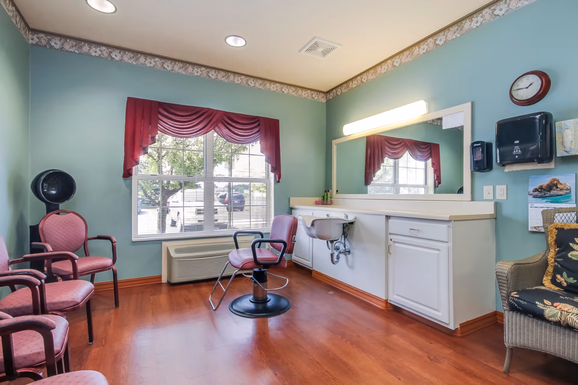 A small salon room with teal walls and wood flooring featuring a single salon chair in front of a large mirror and countertop with a built-in sink. There are three pink cushioned chairs along one wall, a hair dryer hood, a window with red valance curtains, and a wicker chair with floral cushions. A clock and a paper towel dispenser are mounted on the wall.