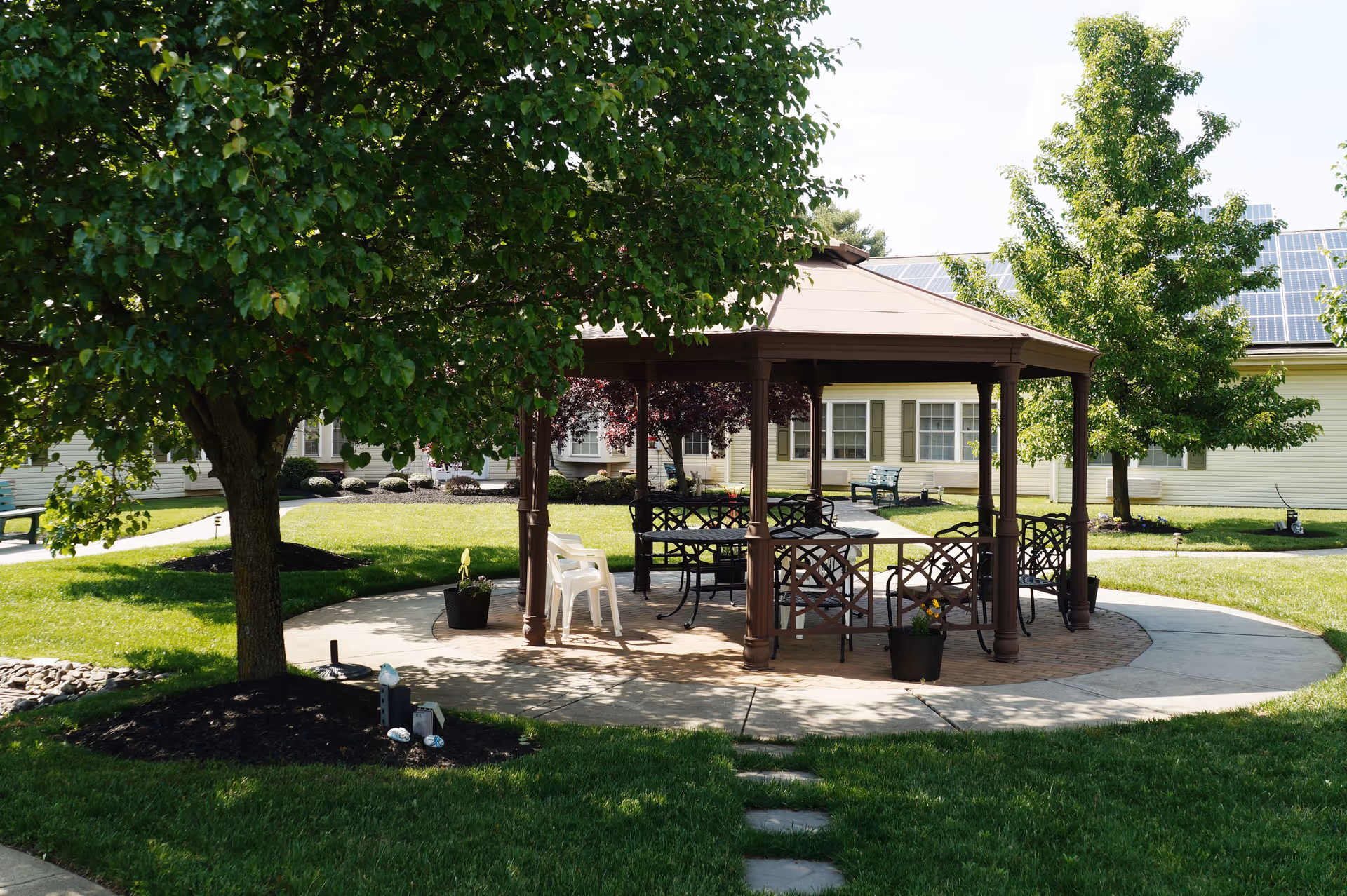 A shaded outdoor gazebo with metal chairs and tables on a circular paved area surrounded by green grass, trees, and a building with solar panels on the roof in the background.