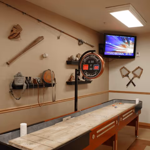 Indoor game room with a shuffleboard table, a digital scoreboard, and a wall-mounted TV. The walls are decorated with vintage sports equipment including baseball bats, gloves, balls, and other memorabilia.