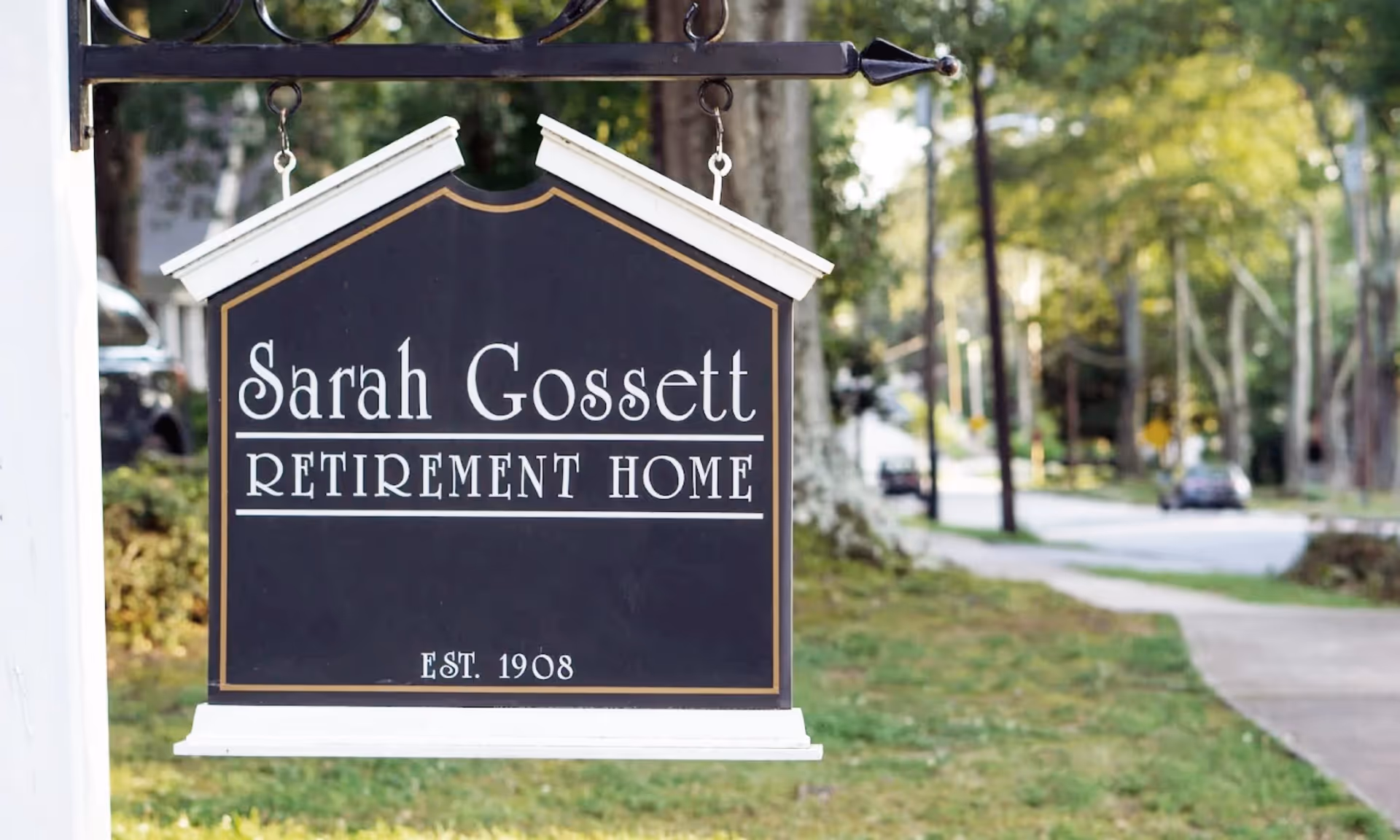 A hanging black sign reads 'Sarah Gossett Retirement Home' in front of a tree-lined street.