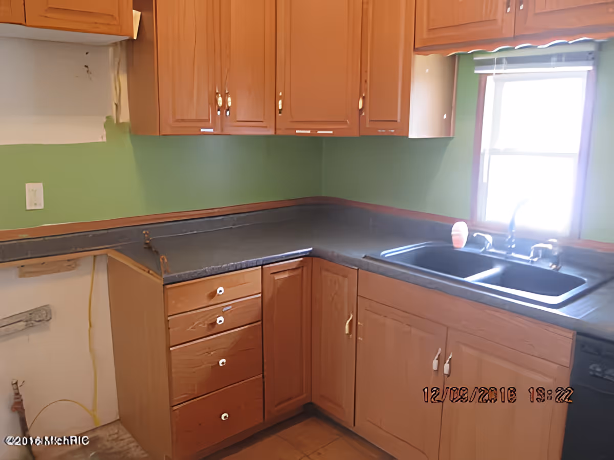 A kitchen corner with wooden cabinets, a black countertop, and a double sink under a window with a partially open blind. The walls are painted green, and there is an empty space where an appliance might be installed. The image has a timestamp in the bottom right corner.