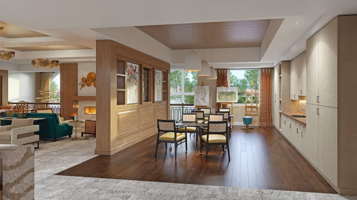 Spacious senior living communal dining and lounge area with a central dining table and chairs, a kitchenette to the right, and a seating area with sofas and a fireplace to the left.