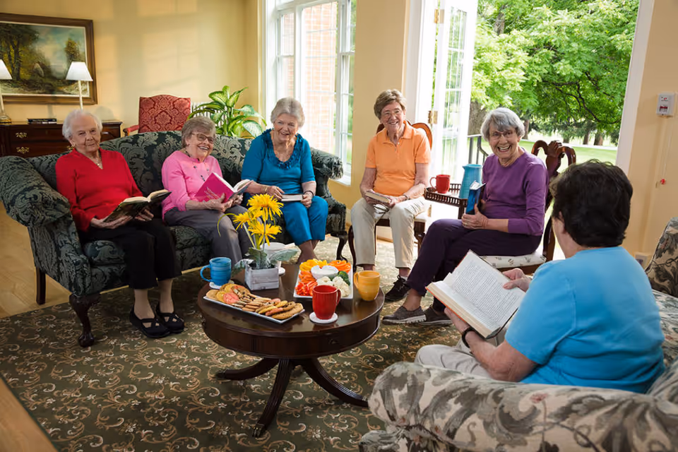 A group of six elderly women sitting in a cozy living room, engaged in reading books and smiling. They are seated on sofas and chairs arranged around a wooden coffee table with snacks, drinks, and a vase of yellow flowers. Large windows and a door open to a green outdoor area, letting in natural light.