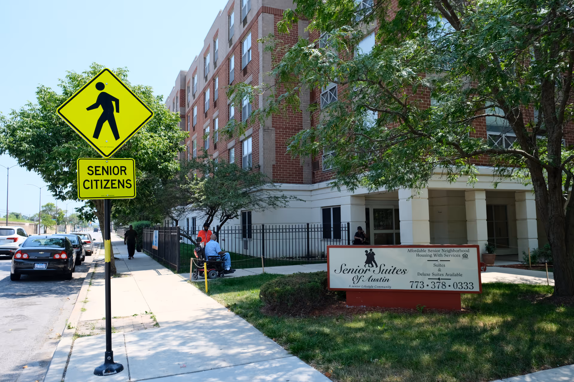 Exterior view of a senior living facility named Senior Suites of Austin. The image shows a multi-story brick and white building with trees and a sidewalk in front. A yellow pedestrian crossing sign with the text 'Senior Citizens' is visible near the sidewalk. Several people are outside the building, including one person in a wheelchair. A sign in front of the building displays the facility name, services, and contact number.