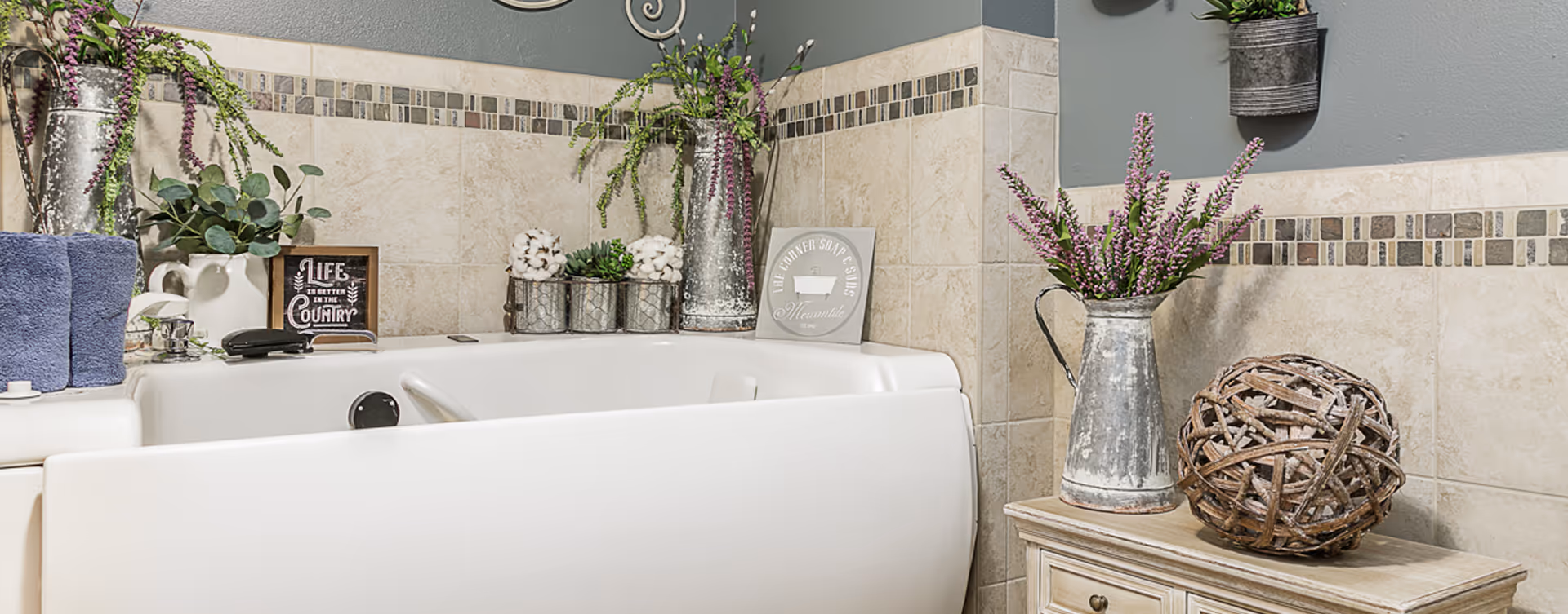 A bathroom corner featuring a white walk-in bathtub surrounded by beige tiled walls with a decorative mosaic border. The bathtub area is decorated with various plants in metal containers, a small framed sign that reads 'Life is better in the country,' and a rustic wooden ball on a small wooden cabinet.