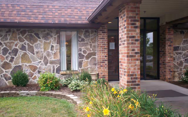 Brick and stone exterior with a covered entrance, glass door and window, and landscaped flowerbeds in front.