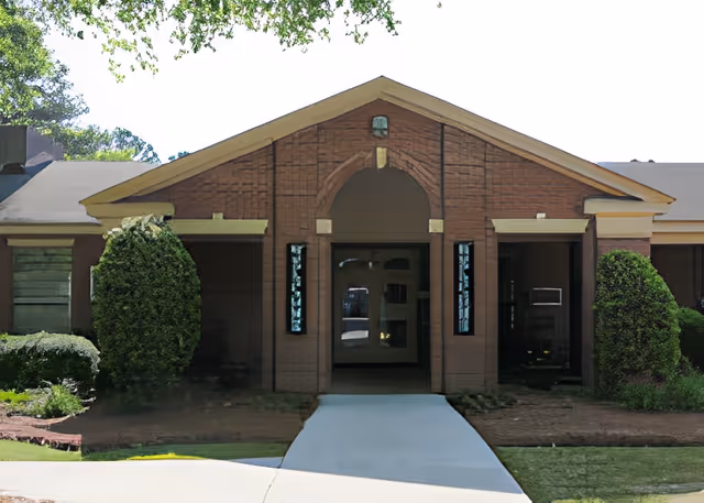 Front exterior view of a single-story brick building with a peaked roof and an arched entrance. There are two tall, narrow windows on either side of the entrance, and trimmed bushes flank the walkway leading to the door. Trees and greenery are visible in the background.