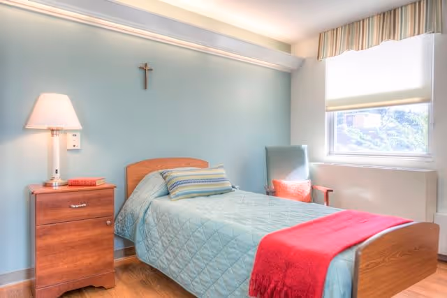 A single bed with a light blue quilt and a red blanket folded at the foot, a striped pillow, a wooden nightstand with a lamp and a book, a green armchair with a red pillow, a window with a striped valance, and a crucifix on the light blue wall.