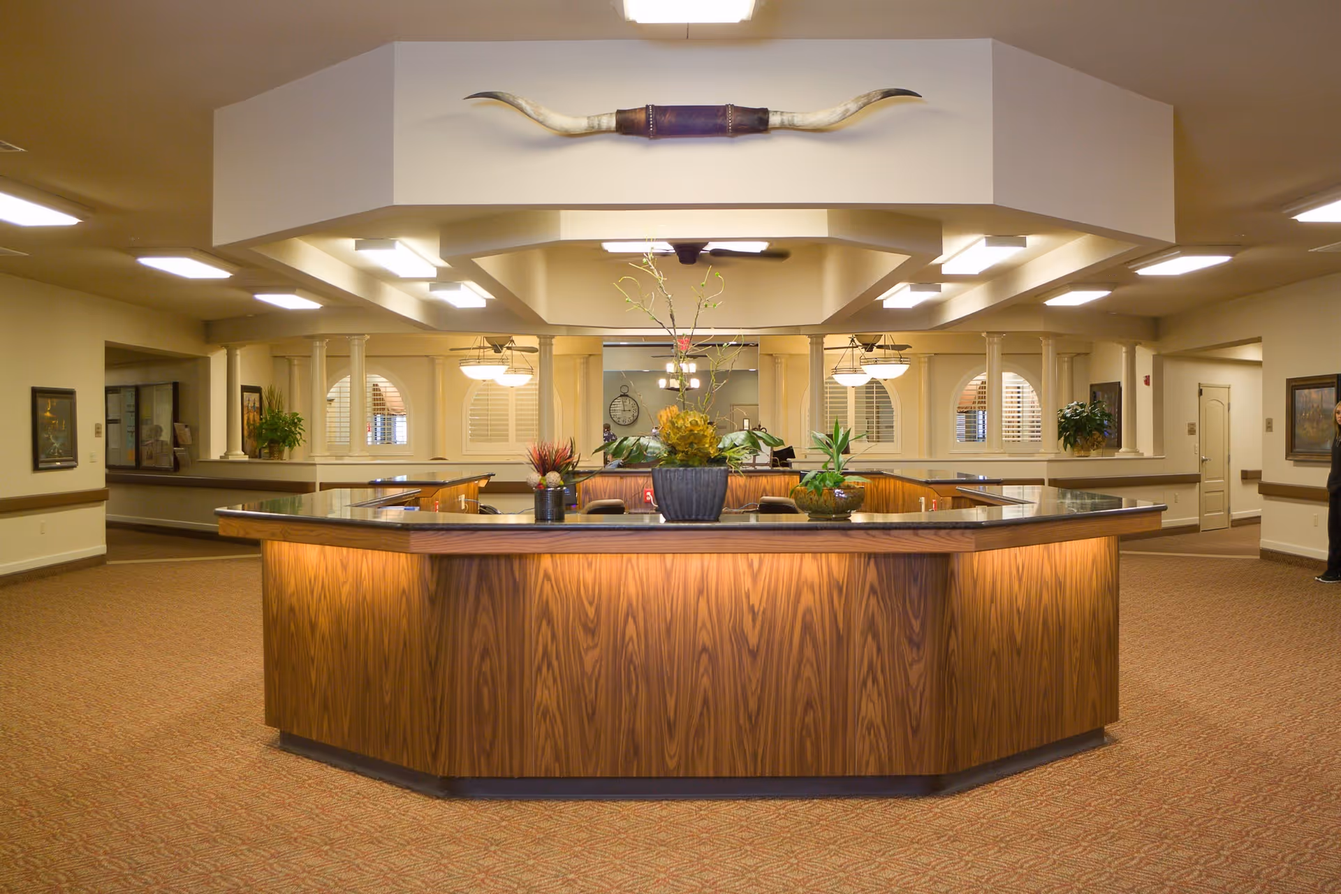 Large, well-lit reception lobby with a central wooden front desk, potted plants, and decorative ceiling accents.