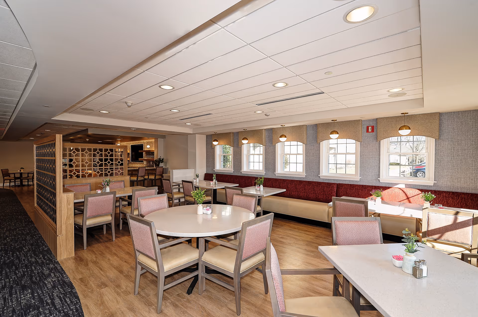 A bright dining room with multiple tables and chairs arranged neatly. The room features large windows with beige valances, allowing natural light to fill the space. There are small plants and condiments on the tables, and a decorative wooden partition separates part of the room. The flooring is wood, and the ceiling has recessed lighting.