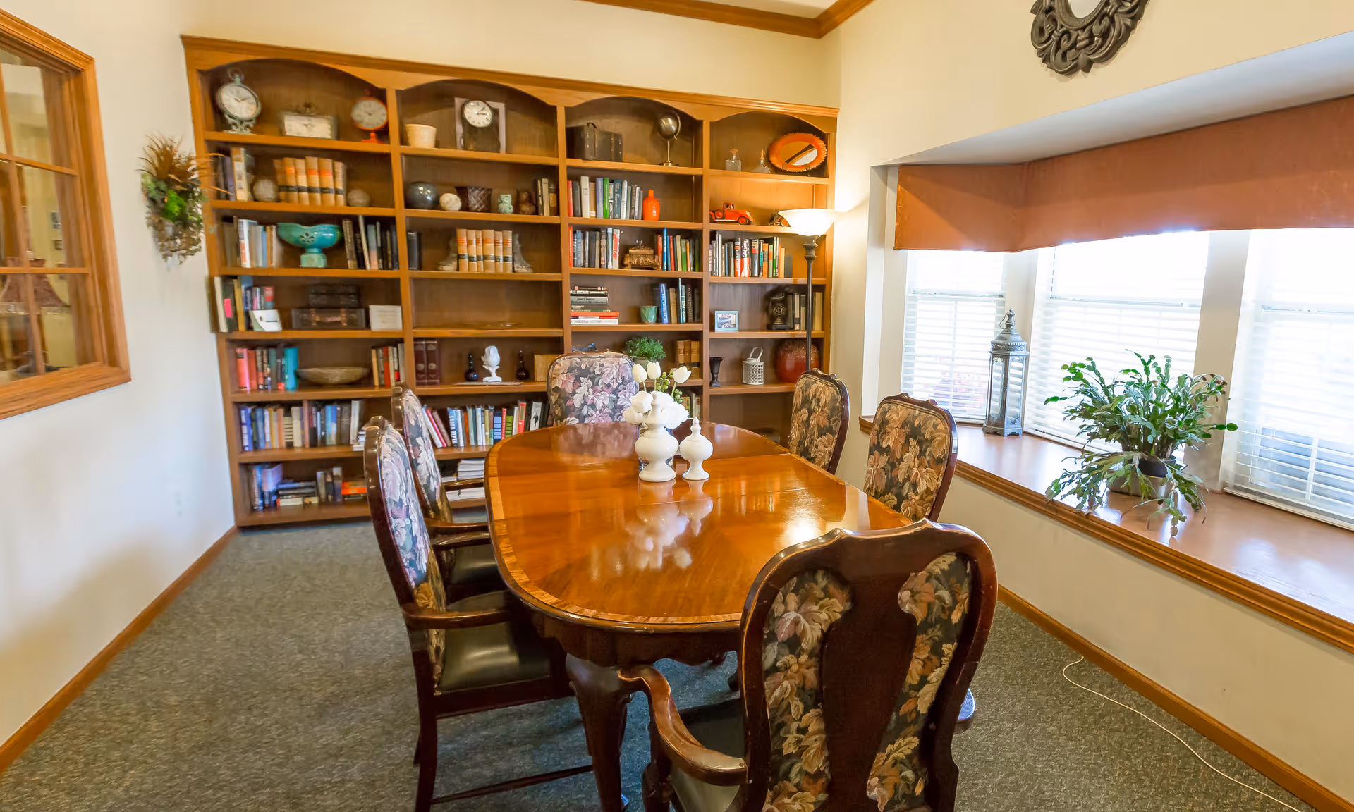 A polished wooden dining table with floral-upholstered chairs in a room featuring a large built-in bookshelf and bright windows with a window seat and plants.