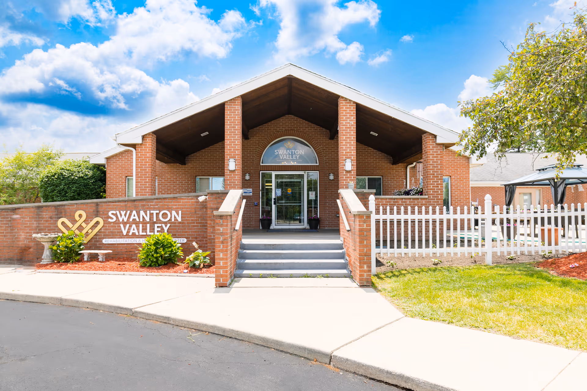 Front exterior view of Swanton Valley Rehabilitation and Healthcare Center, showing a brick building with a covered entrance, steps leading up to glass doors, a white picket fence, and a sign with the facility name on a brick wall. The sky is blue with some clouds.
