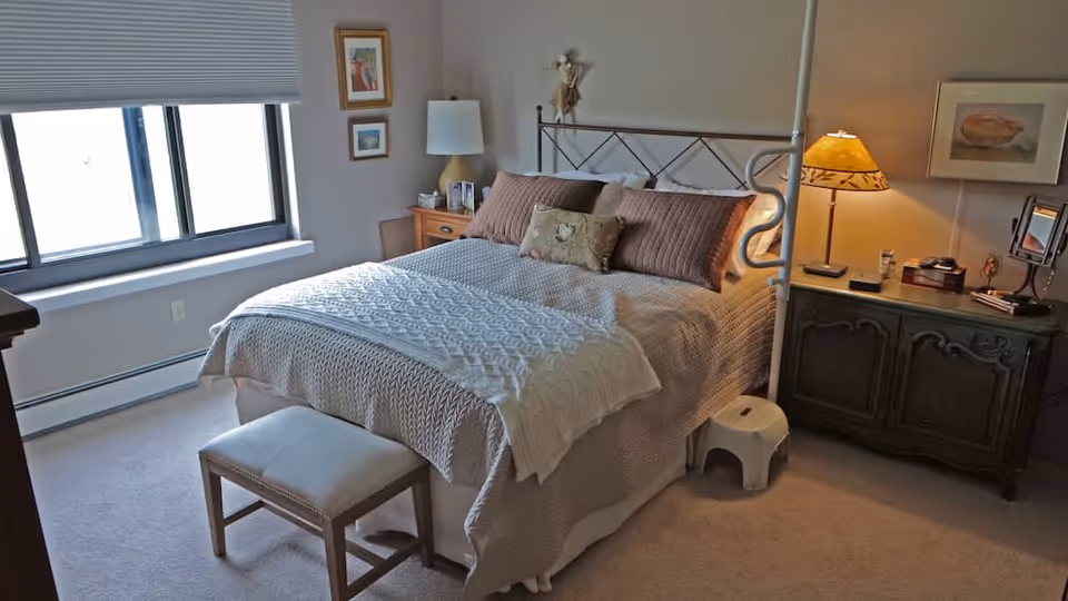 A cozy bedroom with a large bed covered in a beige knitted blanket and decorative pillows. There is a small bench at the foot of the bed, a wooden nightstand with a lamp and picture frames on the left side, and a dark wooden dresser with a lamp, mirror, and other small items on the right side. A window with closed blinds is on the left wall, and framed artwork hangs on the walls.