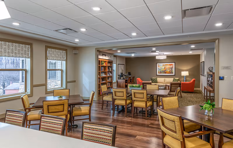 A bright and spacious common area in a senior living facility featuring multiple wooden tables and chairs with yellow cushions. The room has large windows with patterned blinds allowing natural light to enter. In the background, there is a cozy seating area with a beige sofa, two red armchairs, a floor lamp, and a colorful painting on the wall. Bookshelves and a computer workstation are also visible. The flooring is a combination of wood and carpet, and the ceiling has recessed lighting.