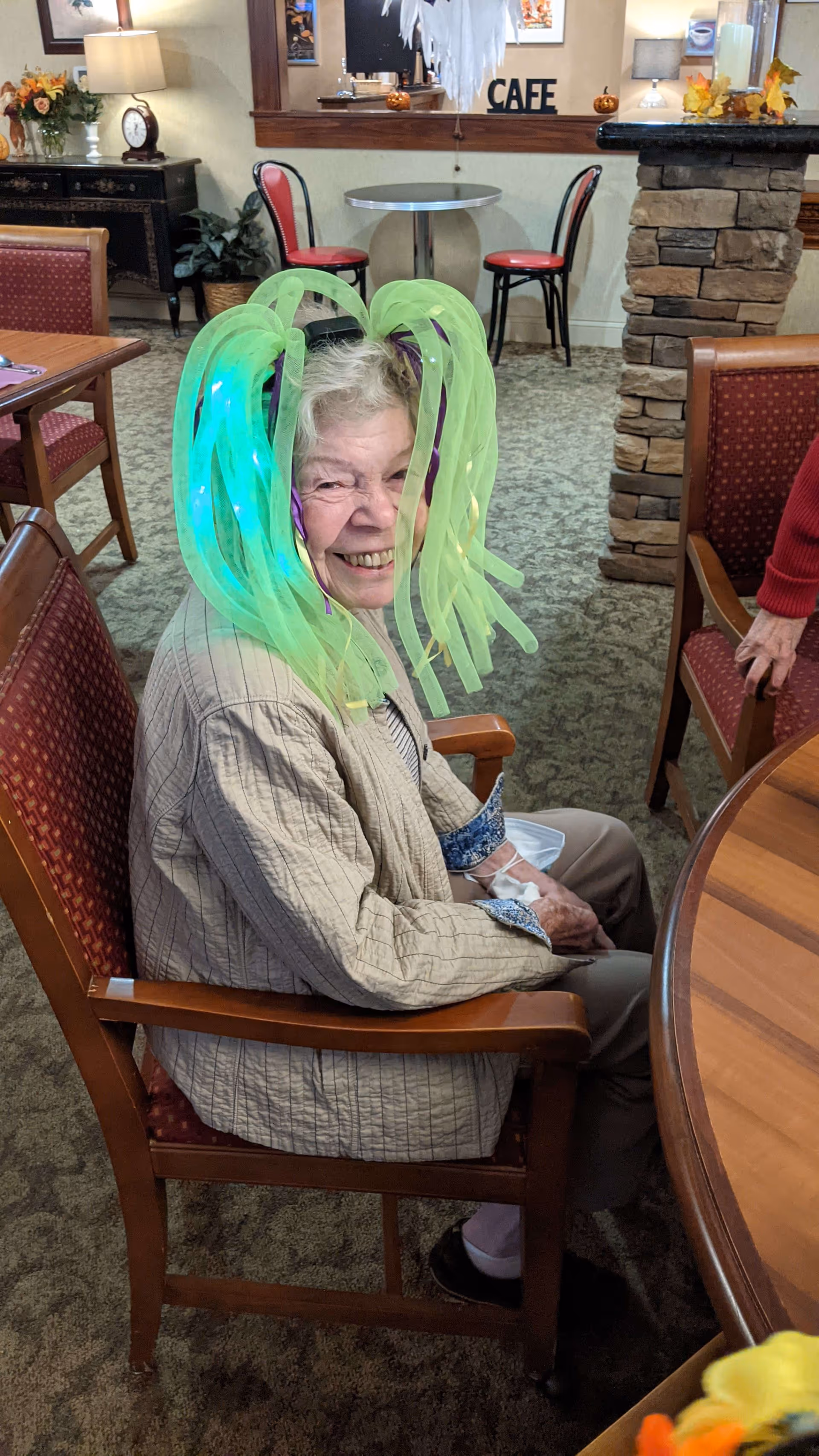 An elderly woman sitting in a chair inside a cozy room, smiling and wearing a glowing green light-up wig. The room has carpeted floors, wooden furniture, and autumn-themed decorations on a stone pillar and a side table. In the background, there is a small round table with two red chairs and a sign that reads 'CAFE'.