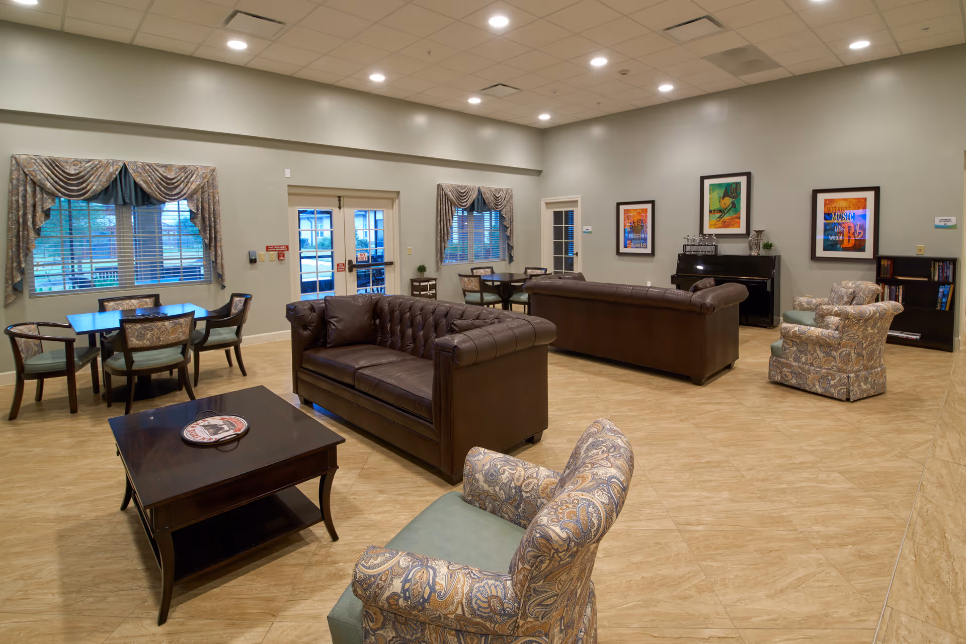 A spacious senior living facility common area with two brown leather sofas, two patterned armchairs, a dark wooden coffee table, and two small dining tables with chairs near windows with draped curtains. The room has beige tiled flooring, light-colored walls, framed artwork, a piano, and a bookshelf.