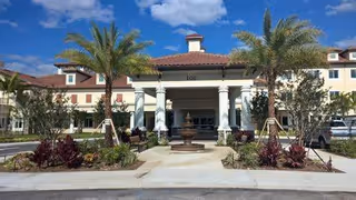Front exterior view of Discovery Village At Palm Beach Gardens building with a covered entrance supported by white columns, a central fountain, palm trees, and landscaped plants under a blue sky with some clouds.